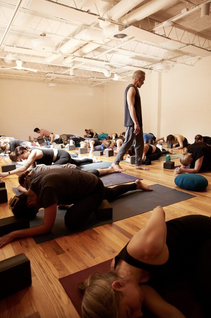 A group yoga class with participants in child’s pose on mats in a bright room. An instructor stands, observing.